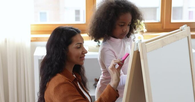 African mom and 6s daughter drawing on white board with colored chalks, family have fun enjoy hobby, communicate in cozy nursery at modern home. Kids development, art class, creative pastime concept