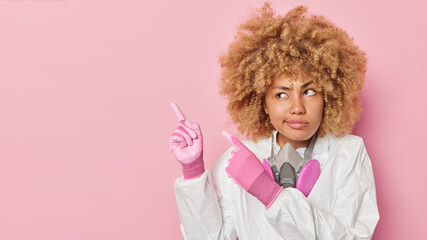 Serious curly haired woman scientist wears protective suit gloves and respirator around neck tells about toxic waste and pollution at ecological disaster site points at blank space against pink wall