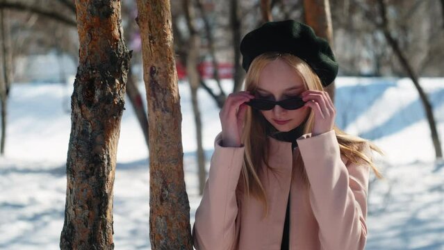 Attractive young model in sunglasses posing in winter forest. Beautiful young woman wearing beret and sunglasses in winter grove on sunny day