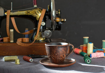 Fototapeta na wymiar A clay mug with coffee on the background of an old vintage sewing machine and sewing accessories in the rays of the setting sun. Coffee time, work break, eco-friendly dishes.