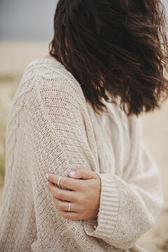 Beautiful woman with windy hair sitting on sandy beach on background of sea, calm tranquil moment. Closeup of stylish young female hand on knitted sweater on cold coast