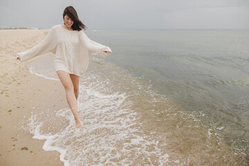 Carefree beautiful woman with windy hair running on sandy beach at cold sea waves, having fun....