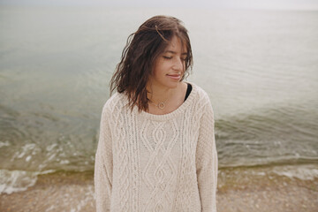 Carefree beautiful woman with curly hair posing on background of  beach and cold sea, tranquil moment. Portrait of stylish young female in knitted sweater relaxing and enjoying day on coast