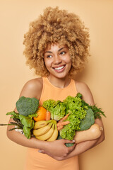 Vertical shot of happy woman poses with fresh green vegetables and fruits keeps to healthy diet...