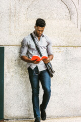 African American Student studying in New York, wearing gray shirt, carrying shoulder leather bag, standing by wall on street, listening music with earphone, reading red book. filtered effect.