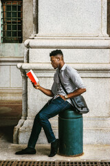 African American College Student studying in New York, wearing gray shirt, jeans, cloth shoes, carrying shoulder leather bag, sitting onl pillar on street, reading red book. filtered effect..