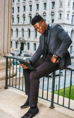 African American Businessman working outside in New York, wearing fashionable jacket, black necktie, sitting on railing in office building, holding tablet computer, reading, thinking, planning.