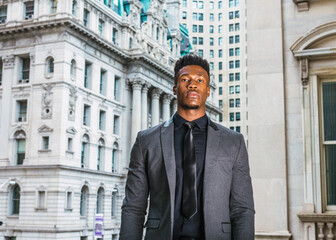 African American Businessman working in New York. Black college student with little goatee, wearing fashionable jacket, black undershirt, necktie, standing by vintage style office buildings on campus.