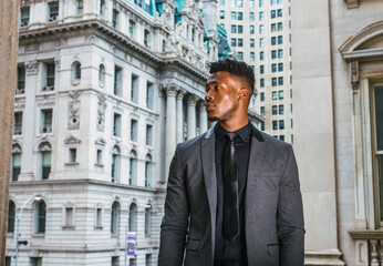 African American Businessman working in New York. Black college student with little goatee, wearing fashionable jacket, black undershirt, necktie, standing by vintage style office buildings on campus.