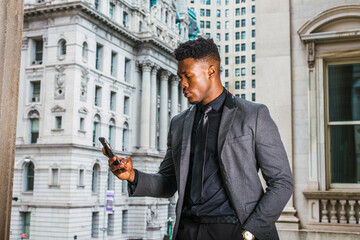 Texting outside. African American Businessman working in New York, wearing fashionable jacket, black necktie, standing by vintage style office building, looking down, reading messages on cell phone.