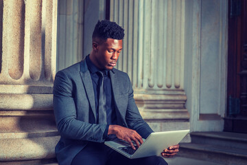 African American Businessman working in New York. Wearing fashionable jacket, necktie, young black man sitting outside office building, typing, working on laptop computer. filtered effect..