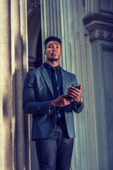 African American Businessman working in New York, wearing fashionable jacket, black necktie, standing by vintage office doorway, reading messages on cell phone, thinking. filtered effect..