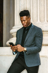 African American Businessman working in New York, wearing fashionable jacket, black necktie, standing by vintage office doorway, reading messages on cell phone, thinking. filtered effect..