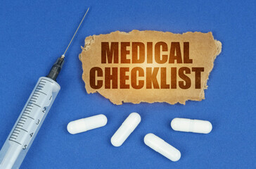 On a blue surface lie a syringe, pills and a cardboard sign with the inscription - MEDICAL CHECKLIST