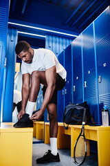 Vertical full length portrait of black sportsman tying shoelaces while changing in locker room...
