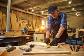 Portrait of a senior carpenter working with a wood, marking plank with a pencil and taking measurements to cut a piece of wood to make a piece of furniture in the carpentry workshop