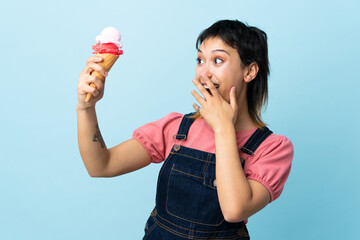 Young Uruguayan girl holding a cornet ice cream over isolated blue background with surprise and...