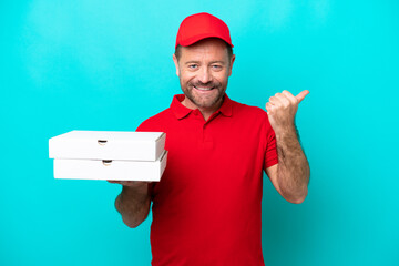 Pizza delivery man with work uniform picking up pizza boxes isolated on blue background pointing to...