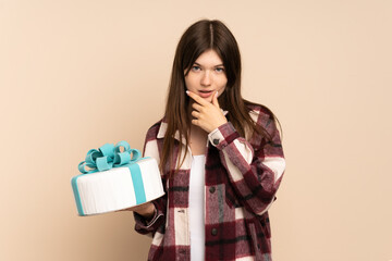 Young Ukrainian girl holding a big cake isolated on beige background thinking