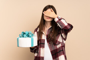 Young Ukrainian girl holding a big cake isolated on beige background covering eyes by hands. Do not want to see something