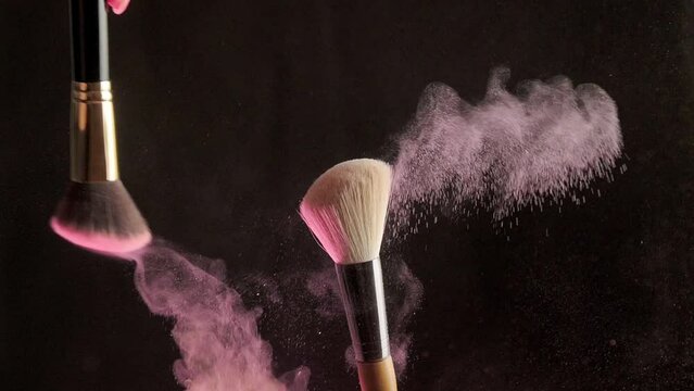 From the touch of two makeup brushes, colorful glittering powder or blush particles float in the air. Bright creative makeup, concept on black background in pink neon light. Slow motion