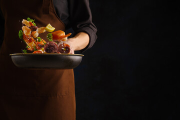 A professional chef prepares seafood - pieces of red fish, shrimp, octopus with vegetables and herbs in a pan on a black background. Advertising, banner. There is free space to insert.