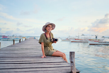 Fototapeta na wymiar Photography and travel. Young woman in hat holding camera sitting on wooden fishing pier with beautiful tropical sea view.