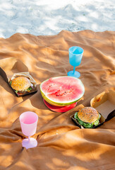 Two burgers and watermelon with glasses on brown cloth on sand