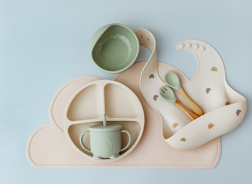 Set of silicone dishware and baby accessories on blue light background, flat lay