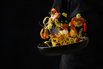 A bright festive dish - a mix of seafood, Italian pasta and vegetables in a frying pan. Explosion of colors and colors. The process of cooking seafood by a professional chef on a black background.