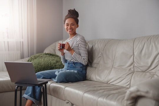 Young beautiful mixed-race female sitting on couch and smiling to camera holding cup of tea