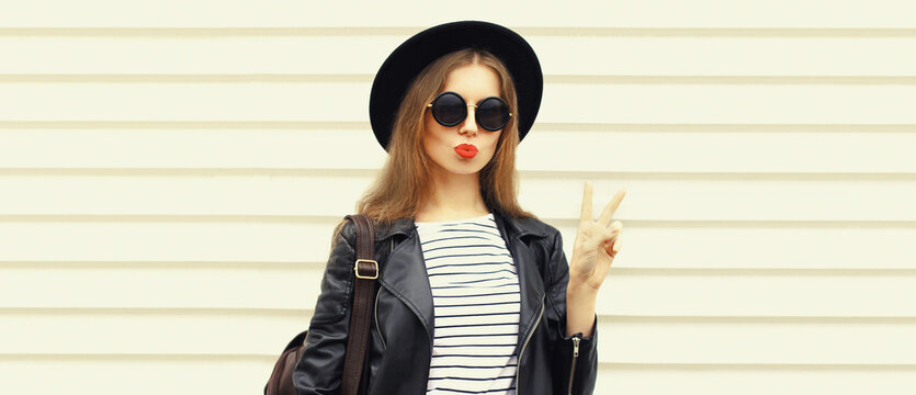 Fashionable portrait of stylish young woman blowing her lips and posing wearing black rock biker jacket, round hat and backpack on white background, blank copy space for advertising text