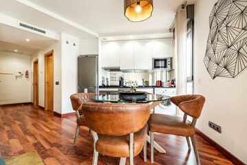 Living room with circular glass dining table with leather and wood chairs, reddish parquet floors and open kitchen with appliances