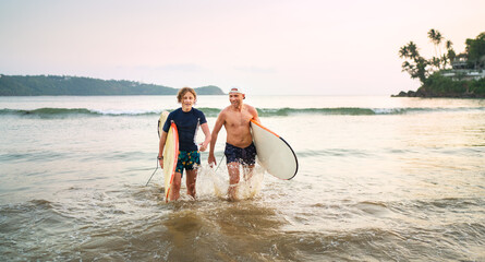 Portrait of a teen boy with his father with surfboards after surfing. They are smiling and walking...