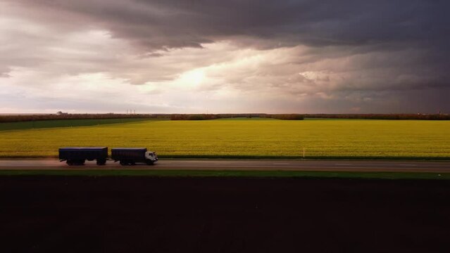 The truck drives along the asphalt highway against the backdrop of beautiful agricultural fields and sunset in the sky. Cargo transportation or commercial delivery at dawn or dusk on the roads