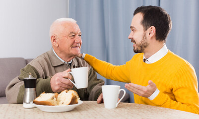 Smiling elderly father and son having breakfast and warmly talk at table at home