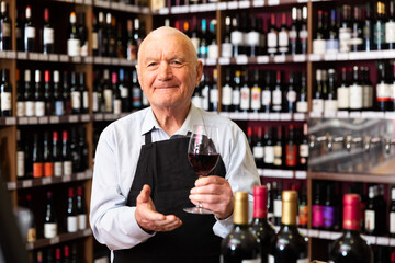 Confident elderly male winemaker inviting to wine house, offering glass of wine for tasting. High...
