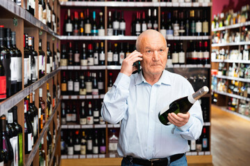 Happy cheerful positive senior man talking on mobile phone and choosing bottle of wine at the liquor store