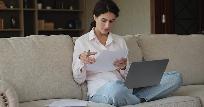 Young woman prepare research or essay sit on sofa with laptop, take notes, make corrections, write up information, do paperwork, report looks serious and concentrated. Tech, freelance workflow concept