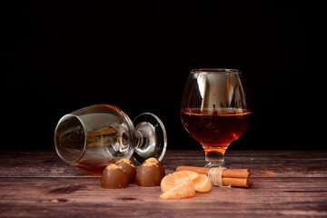 Two glasses of cognac, chocolates, tangerine slices and cinnamon sticks on a wooden table.