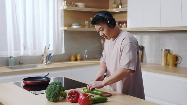 Shooting of cheerful happy Asian guy listening to music in black headphones while cooking dinner in cozy kitchen at home. Vegetables are on the table. Leisure, hobby. Vegetarian lifestyle concept.