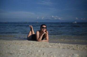 Fototapeta na wymiar A happy woman in glasses and a black swimsuit is lying on the sand. Beach holidays, tourism, vacation and travel. Front view. Sea landscape, blue sky. Philippines beach