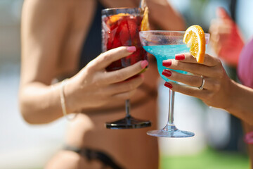 Two tanned female hands clinking with glasses of fresh cocktails, decorated with slice of orange. Crop view of woman hands with manicure holding drinks for toast on vacation. Concept of celebration.