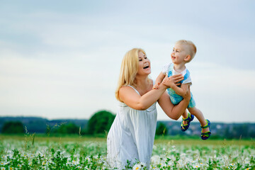 Naklejka na ściany i meble A plump woman and a small one-year-old boy in a field of daisies. A mother holds her son in her arms in a blooming field of camomile.
