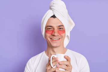 Indoor shot of smiling satisfied man in white towel on head and patches under eyes, holding cup of...