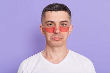 Indoor shot of young adult man with perfect skin wearing moisturizing patches under eyes posing isolated on purple background. Skin care, healthcare cosmetic procedures.