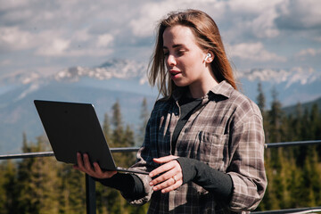A young, slender girl in a plaid shirt works at a laptop in nature against the backdrop of mountains