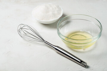ingredients for the French meringue dessert are egg white, sugar and a whisk on a gray surface.