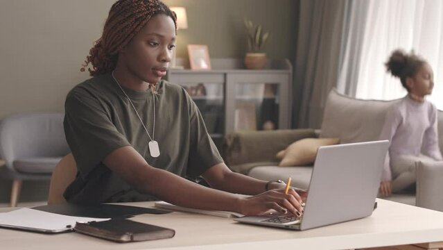 Medium slowmo of concentrated young Black woman soldier working on laptop from home while her playful little daughter jumping on couch in background
