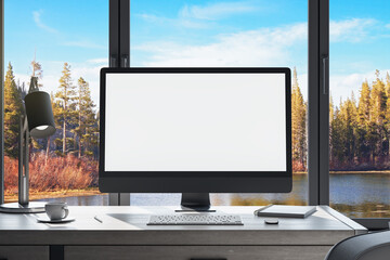 workplace in office interior with blank white mock up computer screen, window with beautiful nature view, coffee cup and other items on desktop, pieces of furniture and daylight. 